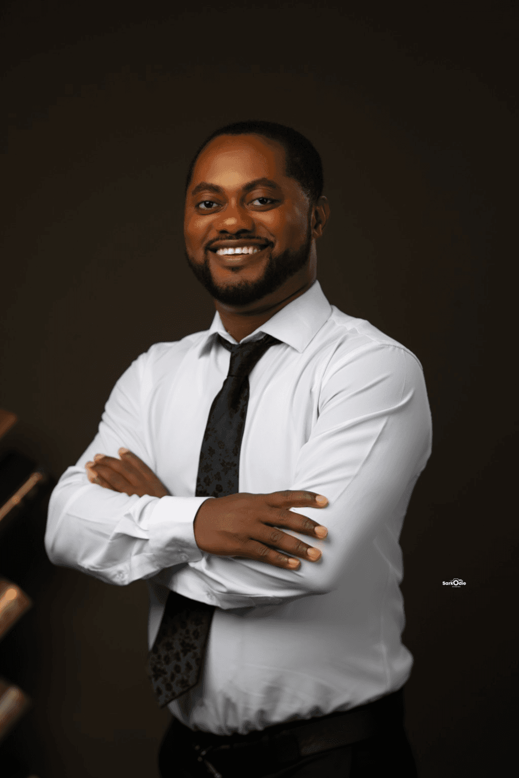Jonah Kwesi Amponsah smiling with arms folded, wearing a white shirt and black tie against the original warm studio background